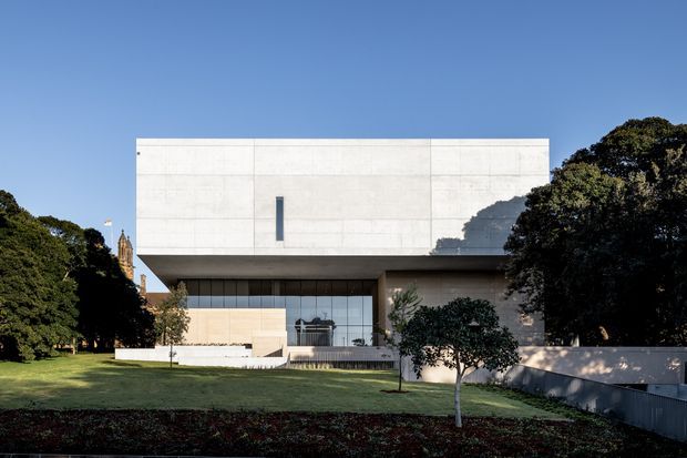A contemporary concrete-clad building photographed from a landscaped lawn, featuring a crisp rectangular upper volume, deep overhang, narrow vertical window, and recessed ground-floor glazing. Mature trees and manicured garden planting frame the minimalist facade.
