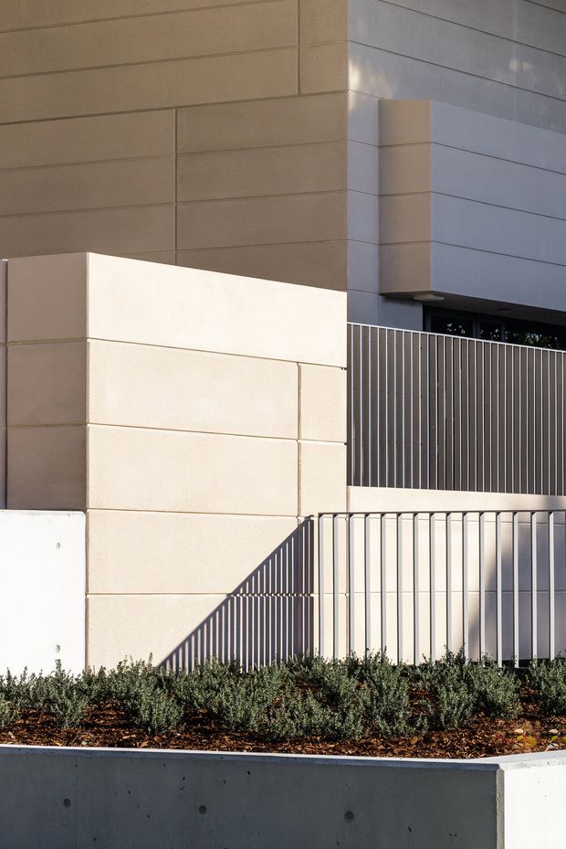 A close-up exterior view of a contemporary building facade featuring large horizontally panelled light beige and grey wall surfaces, projecting box-like volumes, and slim vertical metal fencing. A raised landscaped planter with low shrubs and a concrete retaining wall occupies the foreground. Strong sunlight creates graphic shadows across the facade and fence.