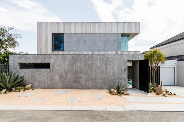 Street-facing view of a contemporary two-storey residence with a monolithic grey concrete ground floor, vertically clad upper level, projecting flat roof, and narrow dark-framed windows. A recessed entry sits beside a white garage door, with sandy paving, stepping stones, and drought-tolerant sculptural planting in the front setback.