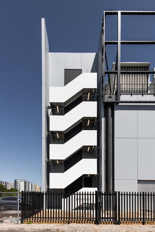 A contemporary multi-storey building exterior featuring a prominent external zigzag stairway in white, dark metal structural framing, grey panelled walls, and a rooftop or upper-level balcony. The building is viewed from outside behind a black metal security fence, with urban buildings visible in the distance under a clear blue sky.