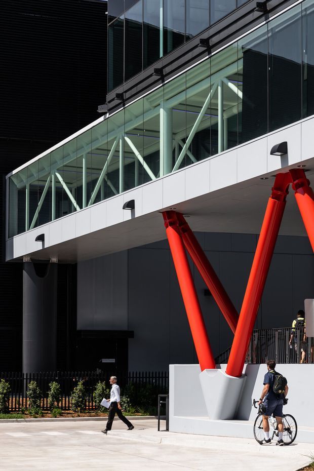 A modern commercial building exterior with a dramatic cantilevered glass walkway or upper-level connector supported by exposed white structural bracing and vivid red diagonal columns. The facade combines dark cladding, extensive glazing, white panelled soffits, and bold structural elements, viewed from a paved public forecourt with pedestrians and a cyclist.