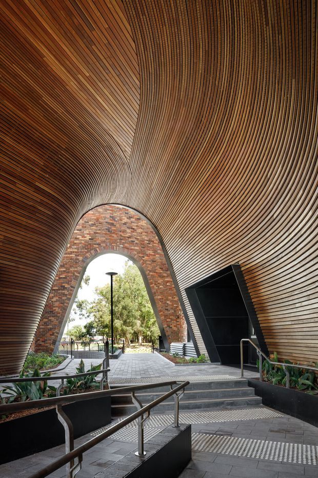 A dramatic public or commercial entry passage with a sweeping, tunnel-like vaulted ceiling clad in closely spaced dark timber slats. A large pointed arch opening is lined with multicoloured brickwork and leads to an outdoor courtyard or landscaped area. The entrance includes dark tiled paving, stairs, ramps, stainless-steel handrails, tactile indicators, integrated planters, and a black angular doorway. The design is highly sculptural and accessible, with strong curved geometry and contrasting timber, brick, and dark metal finishes.