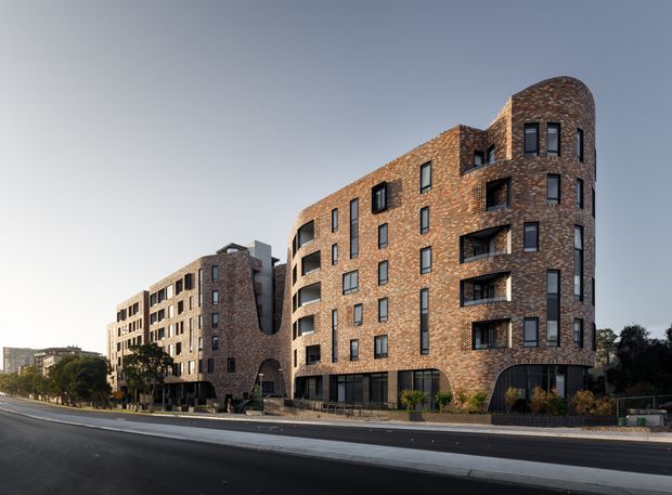 Exterior architectural photograph of a multi-storey apartment building with a curved, rounded corner wing and a connected stepped facade. The building is clad in warm reddish-brown brick with dark-framed windows, recessed balconies, and a central arched opening. It fronts a broad urban street with landscaped planting and neighbouring buildings in the background.