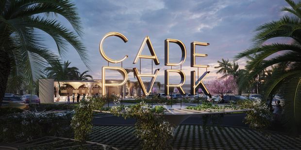 A dusk architectural rendering of a landscaped commercial development entrance, framed by tall palm trees and planting. Large illuminated gold lettering spelling “CADE PARK” forms the prominent gateway signage, with a paved forecourt, parked cars, pedestrians, low modern buildings, and a softly lit purple-blue evening sky in the background.