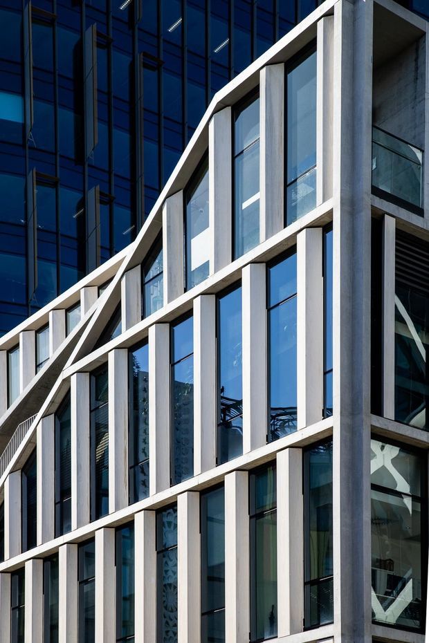 Close-up exterior view of a contemporary multi-storey commercial building facade. The composition features a repeating grid of tall blue-tinted glazed windows framed by pale concrete vertical fins and horizontal bands, with a dramatic diagonal stepped roofline or facade edge. Dark glazed building volumes and visible interior structural elements appear behind the main facade.