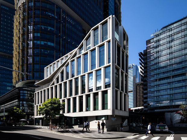 Urban architectural exterior of a contemporary multi-storey building with a distinctive white structural grid facade, tall vertical glazing, and a sloping stepped roofline. The building is set within a dense central business district surrounded by taller glass-and-steel towers, with pedestrians, bicycles, and a street intersection in the foreground.