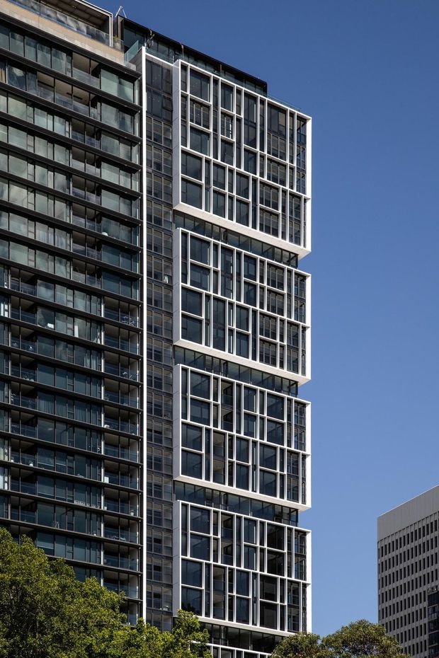 Exterior architectural photograph of a tall contemporary apartment building with a dark glazed facade, extensive balconies, and projecting stacked bays framed by white geometric window surrounds. Trees and a neighbouring office building are visible at the base and edge of the composition against a clear blue sky.