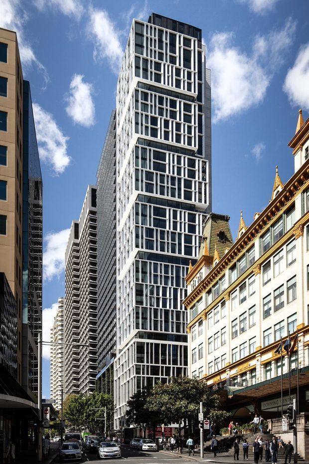 Street-level view of a dense urban city centre, dominated by a tall contemporary high-rise with a strongly gridded glazed facade and projecting white structural frames. Historic ornate masonry architecture with decorative rooflines and gold-toned trim occupies the foreground at right, contrasting with surrounding modern towers. Vehicles, pedestrians, street trees and a bright blue sky are visible.
