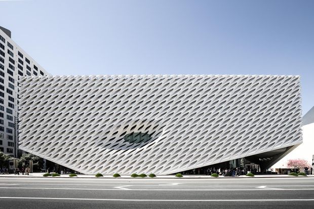 Exterior architectural photograph of a striking contemporary building with a sweeping, perforated white lattice facade forming a sculptural geometric envelope. The building sits prominently along a broad urban street, with taller modern buildings visible behind it. The image emphasizes the facade, unusual structural form, and urban setting.