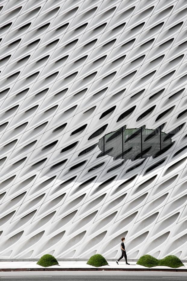Close-up exterior view of a striking contemporary building facade with a repetitive white sculptural lattice of curved, elongated openings. Dark glazing is integrated into the patterned envelope, with a pedestrian walkway and neatly clipped shrubs at street level.