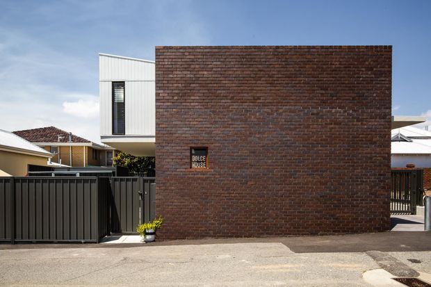 Street-facing view of a contemporary urban residence, dominated by a dark red-brown brick wall with a small “Dolce House” plaque. A black metal gate and fencing sit in front, with a white corrugated upper-level volume and neighbouring residential roofs visible around the masonry facade.