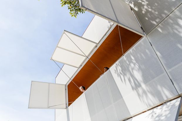 Upward-looking exterior view of a contemporary building with crisp white geometric cladding and projecting folded or cantilevered panels. A recessed opening reveals a warm timber-lined soffit or ceiling, with two people visible within the opening. Strong angular forms, deep overhangs, and dappled tree shadows emphasize the sculptural facade.