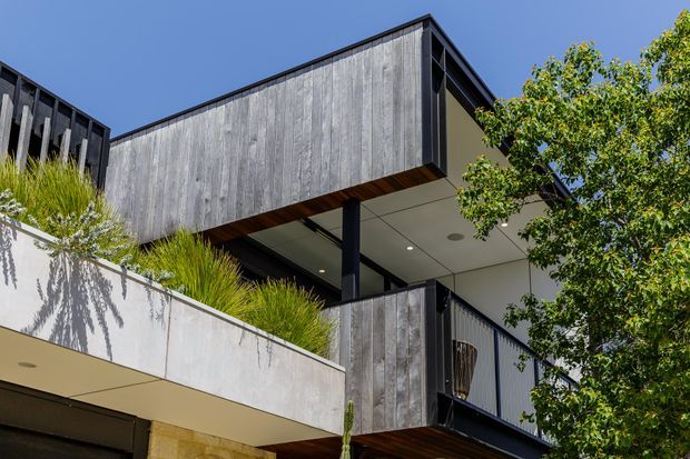 Contemporary multi-level residential exterior with strong cantilevered forms, dark metal framing, weathered vertical timber cladding, pale rendered concrete balcony surfaces, and landscaped planter beds. The image looks upward toward a covered balcony or terrace beneath a crisp flat roofline.