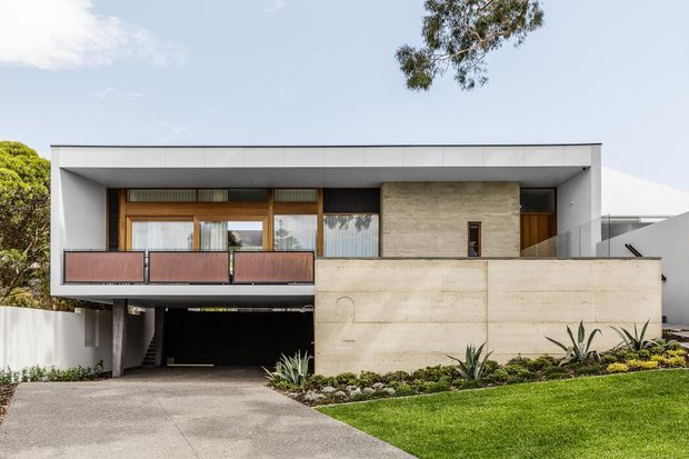 Exterior view of a contemporary two-storey residence with a strong horizontal, flat-roofed form. The facade combines smooth white rendered surfaces, exposed concrete walls, warm timber-framed glazing, and a dark recessed garage or carport beneath the upper level. A planted front garden with architectural succulents borders the concrete driveway.