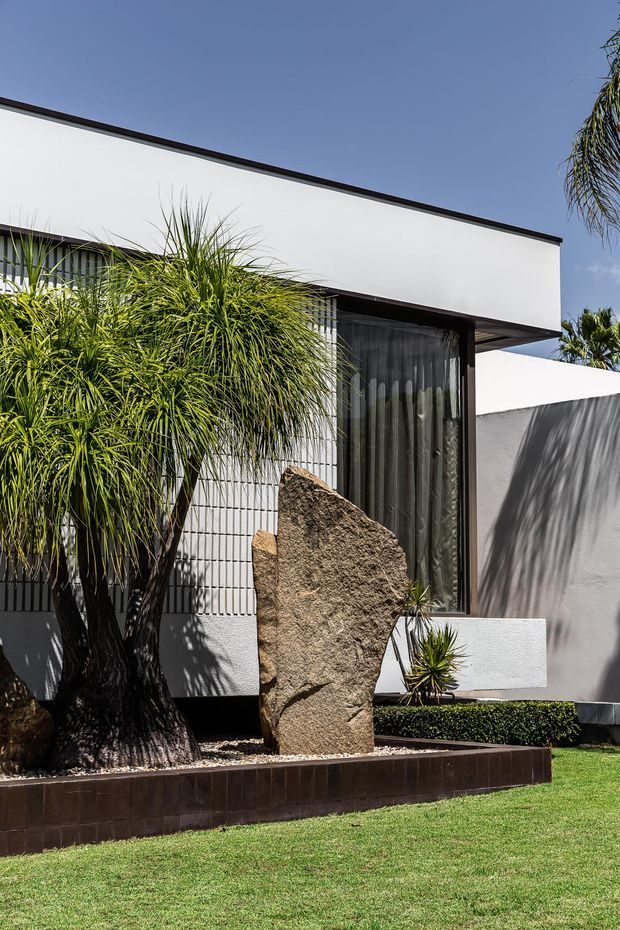 Photograph of a modern, minimalist residential facade with crisp white rendered walls, dark trim, vertical glazing, and a projecting flat roof. The foreground features a manicured lawn, sculptural natural stone, clipped hedging, and tropical-style planting.