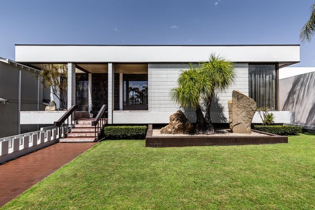 Street-facing view of a low, single-storey modern residence with a crisp flat roof, white rendered facade, dark-framed glazing, a screened entry porch, and a landscaped front garden featuring palms, rocks, hedges, and lawn. A ramp and steps lead to the covered entrance.