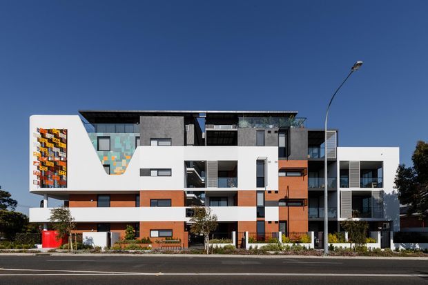 A contemporary multi-storey apartment building viewed from across a street. The facade combines crisp white rendered volumes with dark grey panels, warm orange-brown cladding, extensive glazing, recessed balconies, and a colourful patterned feature wall at the left. Low landscaping and street elements sit in front of the building beneath a clear blue sky.