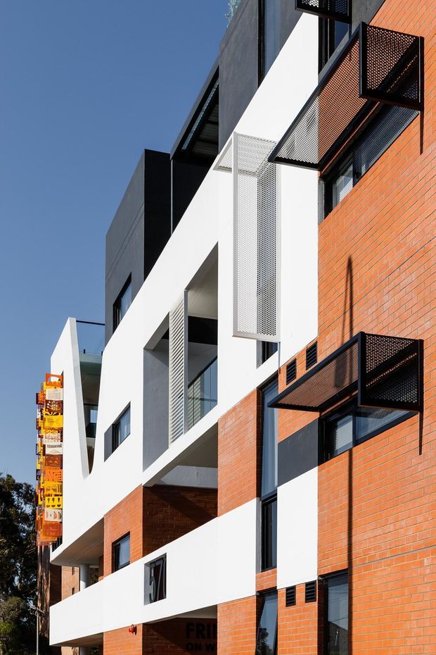 Exterior architectural photograph of a contemporary multi-storey urban building. The facade combines red brickwork, white rendered walls, dark charcoal panels, black-framed glazing, and projecting perforated metal screens and balconies. Strong geometric massing and recessed openings are visible against a clear blue sky.