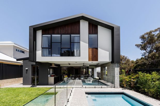 Front exterior view of a contemporary multi-level residence with a dramatic gabled roof, dark framed facade, white rendered walls, timber feature cladding, extensive glazing, and an open ground-level entertaining area. A swimming pool, glass balustrades, landscaped garden, and adjacent outdoor living space are visible.