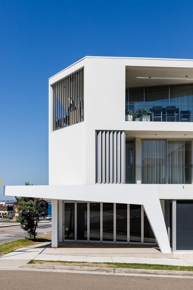 Street-facing view of a contemporary white multi-storey building with clean rectangular volumes, deep balcony recesses, extensive glazing, and vertical sun-screen fins. The ground level appears commercial or office-like, while upper levels contain balconies with outdoor furniture. The building occupies an urban or suburban streetscape under a clear blue sky.