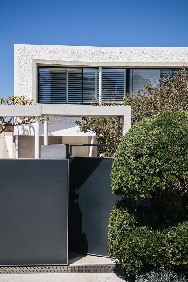 Exterior view of a contemporary two-storey residence with a white textured rendered facade, broad horizontal louvred windows, a concrete-framed entry canopy, dark metal pedestrian gates, and carefully clipped shrubs in the foreground.