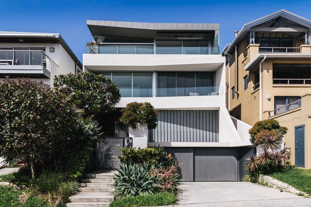 Street-facing view of a contemporary multi-storey residence with a white rendered facade, broad glazed openings, recessed balconies, a projecting flat roof, and a dark vertical-slat garage door. The home is set within a landscaped suburban frontage and flanked by neighbouring residences.