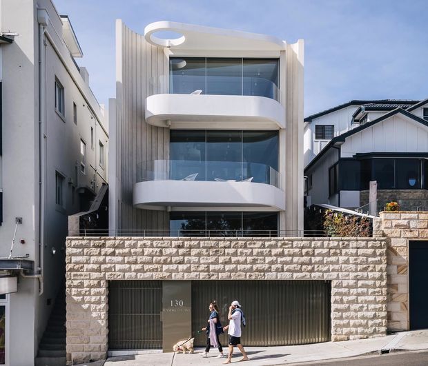Street-facing view of a narrow multi-storey contemporary residence with a sculptural white facade, curved projecting balconies, extensive floor-to-ceiling glazing, vertical ribbed wall cladding, and a large integrated garage. A pale textured stone boundary wall fronts the property, with neighbouring homes closely flanking both sides.