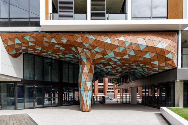 Contemporary multi-storey building entrance with a dramatic cantilevered canopy and supporting column clad in a colourful geometric mosaic of triangular copper, terracotta, cream, grey and turquoise panels. Extensive glazing, white projecting volumes, louvred openings and dark framed storefront-like entrances surround a paved forecourt.