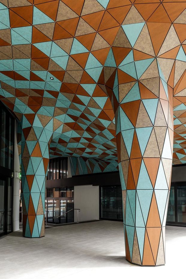 A striking contemporary interior atrium or lobby with large faceted structural columns and a ceiling clad in an irregular geometric mosaic of terracotta, turquoise, beige, and dark brown panels. Full-height glazing, dark framed doors, a mezzanine-like internal level, and a pale tiled floor create a dramatic public or commercial space.