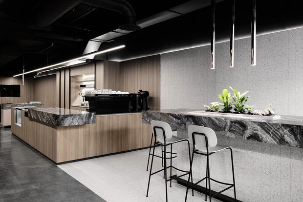 A sophisticated contemporary commercial café or hospitality interior with a long stone-clad counter and integrated seating. The space features dark exposed services and ceiling elements, linear architectural lighting, warm timber cabinetry, grey textured wall finishes, and dramatic veined stone benchtops. A central island supports a professional espresso machine, while minimalist black-framed bar stools sit at the counter.