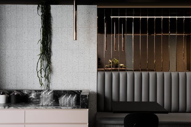 A moody, refined hospitality-style interior featuring a dark dining or lounge booth with vertically padded upholstery, a black table, and a dramatic arrangement of slender pendant lights. A pale stone or concrete partition separates the booth from a compact refreshment counter with dark veined stone, pale cabinetry, glassware, and greenery. The space uses strong vertical lines, reflective surfaces, and integrated lighting.