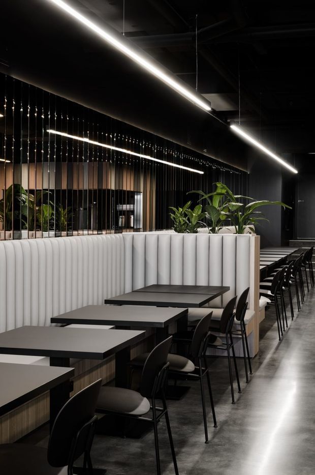 A contemporary commercial dining interior with rows of black tables and chairs beside a long white vertically channelled upholstered banquette. The space features dark walls and ceiling, polished concrete flooring, suspended linear LED lighting, black metal detailing, timber-toned panels, and integrated planters with greenery. Reflective vertical rods create a screen and add depth to the restaurant or hospitality setting.
