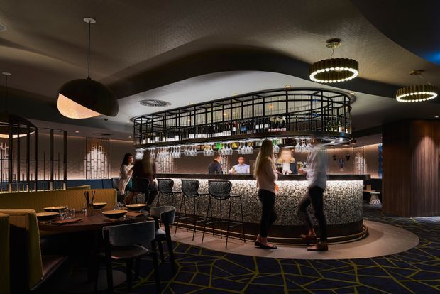 A sophisticated, dimly lit restaurant or cocktail bar interior featuring a curved illuminated bar with a textured stone-like front, high black metal bar shelving, hanging glassware, and backlit bottle displays. The space includes dark dining tables and upholstered chairs, patterned carpet, sculptural pendant lighting, and warm ambient illumination.