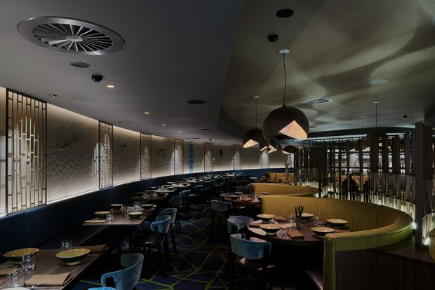 A dimly lit upscale restaurant dining room with curved banquette seating, dark tables, blue upholstered chairs, decorative metal screens, textured illuminated wall panels, pendant lights, and patterned carpet. The space has a long, softly curving layout with an intimate, refined atmosphere.