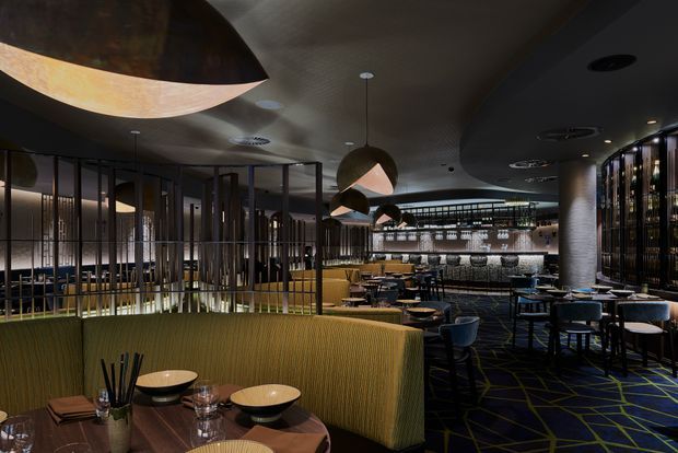 A moody, sophisticated restaurant interior with a bar and dining area. The space features curved olive-green upholstered banquette seating, small round dining tables, dark blue chairs, black-framed glazed partitions, a textured dark ceiling, sculptural pendant lights, a backlit bar, and a dark patterned carpet with geometric yellow lines.
