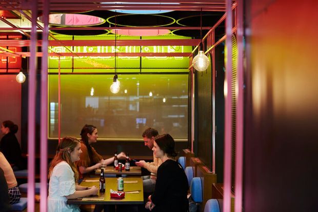 A brightly coloured contemporary commercial dining space, likely a restaurant or casual eatery, with diners seated at compact tables. The interior features vivid pink, red, yellow and lime-green structural frames and translucent partitions, exposed ceiling elements, and hanging spherical pendant lights. Reflections and blurred signage appear on the glazed rear partition.