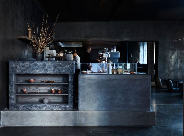 Dark, moody industrial-style commercial interior showing a cafe or bar service counter. The foreground features a large charcoal-grey textured counter with open display shelving holding ceramics and objects, while staff work behind a glazed service counter. Concrete-like walls, dark flooring, exposed dark ceiling elements, and black-framed glazing reinforce the raw contemporary atmosphere. A dim seating area is visible to the right.