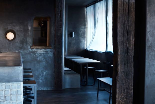 A dark, compact industrial-style interior with rough textured charcoal walls, exposed dark timber posts, concrete built-in bench seating, and small black tables and stools arranged beside a tall curtained window. A recessed shelf and circular wall light add subtle functional details. The space feels intimate and moody, resembling a café, bar, or compact dining area.