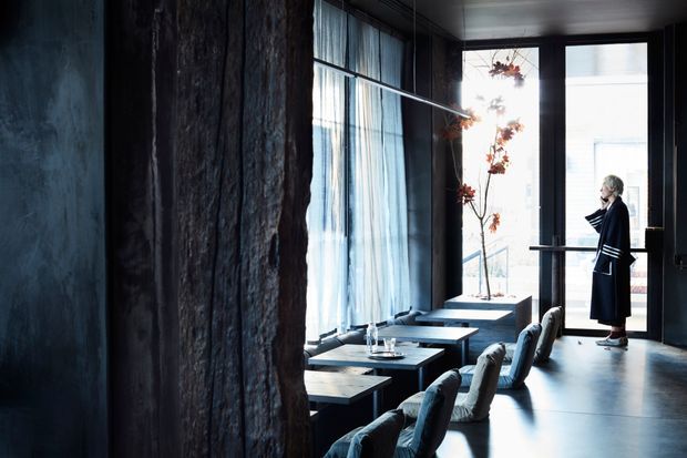 A moody, refined commercial interior with a long row of tables and upholstered chairs beside tall sheer-curtained windows. Dark textured wall surfaces, black-framed glazing, suspended linear lighting, and a polished dark floor create a dramatic hospitality or dining setting. A person stands near the bright glazed entrance at the far end.