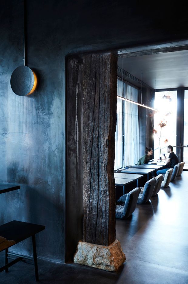 Dark, atmospheric commercial dining interior viewed through a rustic timber-and-stone structural opening. A textured blue-grey wall with a round wall light occupies the foreground, while the adjoining space contains long communal tables, upholstered lounge chairs, pendant lighting, and tall windows with sheer curtains. The palette is moody and industrial with strong natural timber and polished dark flooring.