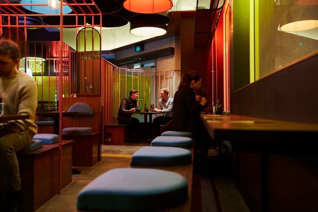 A dimly lit contemporary commercial dining or hospitality interior with booth seating, a long banquette, small tables, and patrons. The space features vivid red and orange metal framing, colourful translucent or painted partitions, dark timber-toned wall surfaces, and multiple oversized circular pendant lights. The atmosphere is intimate, warm, and dramatic, with a strongly graphic, playful fit-out.