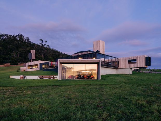 Wide dusk exterior view of a contemporary multi-level building set within a broad grassy landscape. The composition features rectilinear timber-clad volumes, dark-framed floor-to-ceiling glazing, exposed concrete foundations, a rooftop terrace or balcony, and a prominently illuminated glazed living or dining space. Woodland rises behind the building, with outdoor benches and landscaped lawn in the foreground.