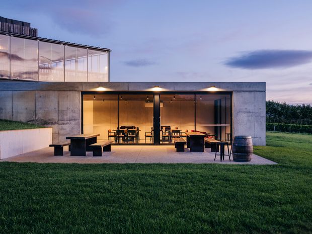A modern single-storey concrete pavilion with extensive floor-to-ceiling glazing, photographed at dusk. The illuminated interior contains a dining area and seating, while the outdoor terrace has dark benches, tables, stools and a barrel. A glazed upper volume projects above the flat-roofed concrete structure, with a broad lawn and open rural-looking landscape surrounding the building.