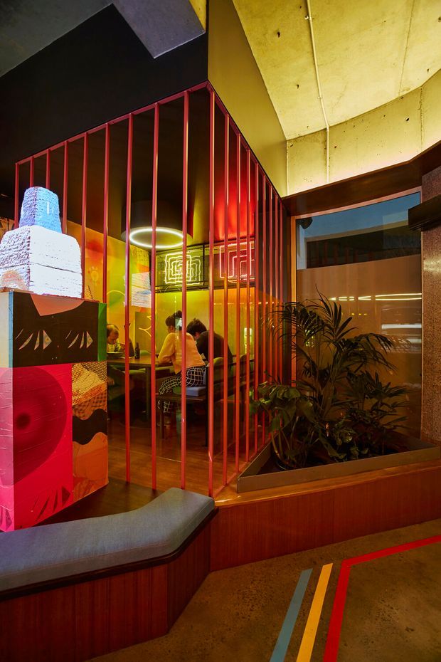 A richly coloured commercial interior with a small dining or lounge area enclosed by tall red metal vertical bars. The space features dark timber wall panelling and bench seating, polished concrete flooring with colourful painted line markings, exposed concrete ceiling surfaces, indoor planting, geometric neon-style lighting, and bold graphic artwork. Diners are visible behind the partition, suggesting a restaurant or hospitality venue with an industrial-retro atmosphere.