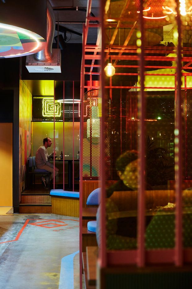 A dimly lit, highly stylised commercial interior with vivid red, orange, yellow and green lighting. The space features booth seating, perforated metal screens, exposed ductwork and ceiling structure, translucent and reflective partitions, patterned concrete flooring, and colourful sculptural lighting. A person is seated at a small table in the background, suggesting a bar, restaurant or entertainment venue.