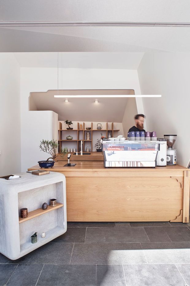 A bright, minimalist café interior focused on a timber service counter with a commercial espresso machine. The space features pale plaster walls, dark large-format floor tiles, warm timber cabinetry and shelving, a sculptural pale stone or concrete display unit, simple linear pendant lighting, and understated natural decor.