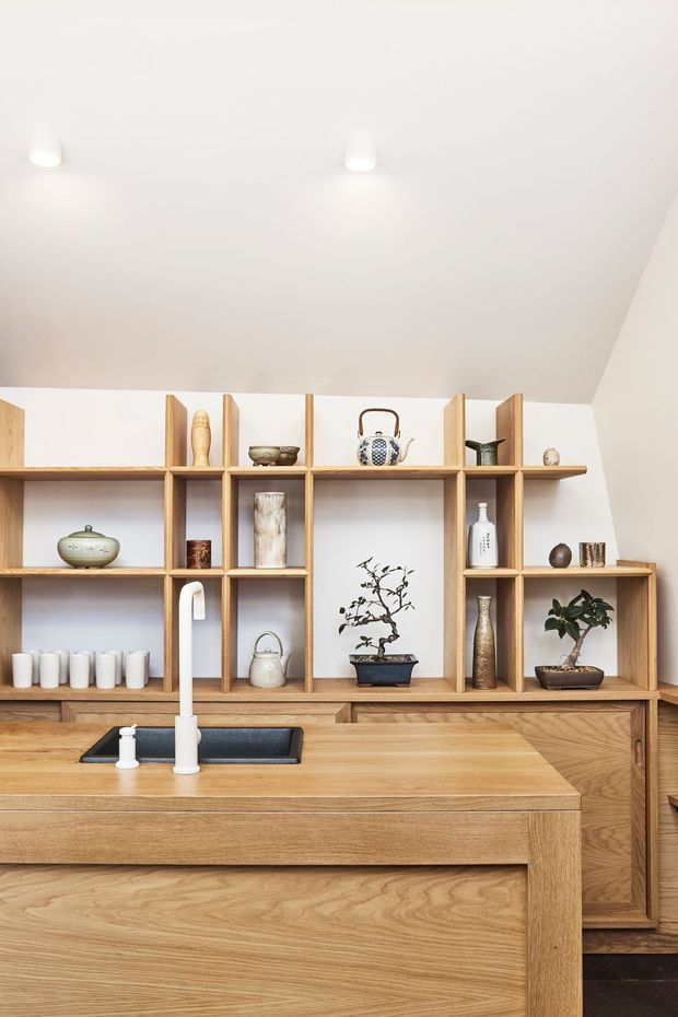 A warm, minimalist kitchen featuring extensive light-to-mid timber cabinetry and an island with an inset dark sink and tall white tap. Open timber shelving displays ceramics, bottles, a kettle, decorative objects, and bonsai plants. The room has white walls and ceiling with recessed downlights and a sloped ceiling profile.