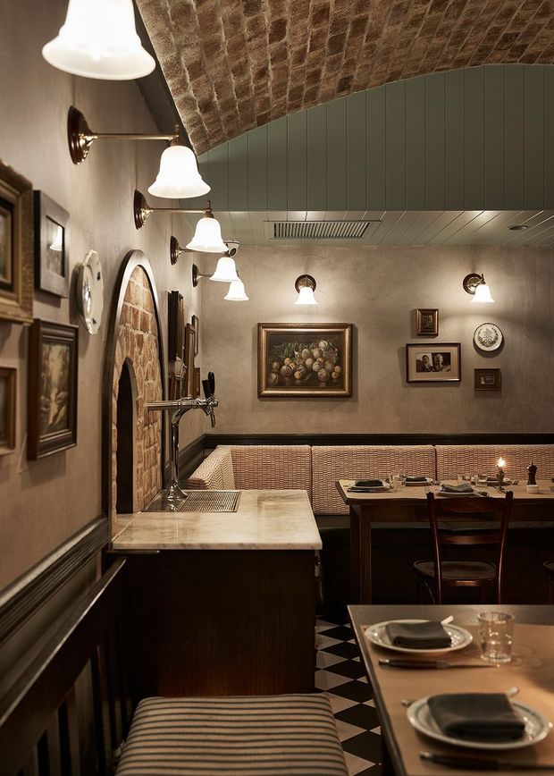 A warmly lit, intimate restaurant interior with a compact bar and marble counter in the foreground, upholstered banquette seating, dark timber dining tables, framed artwork, wall sconces, and a striking curved exposed brick ceiling with a brick arched opening. The space has a refined vintage European character with a black-and-white checkerboard floor and low, atmospheric lighting.