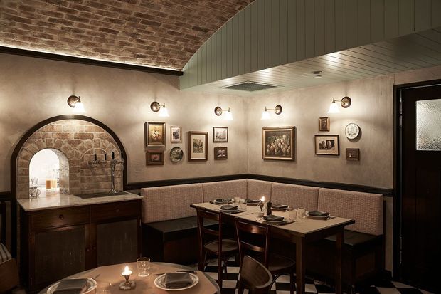 A warmly lit, intimate restaurant dining area with built-in upholstered banquette seating, dark timber dining tables and chairs, framed artwork, exposed brickwork, and an arched brick alcove with a service counter. The space has a refined, historic character with dark finishes and decorative wall lighting.