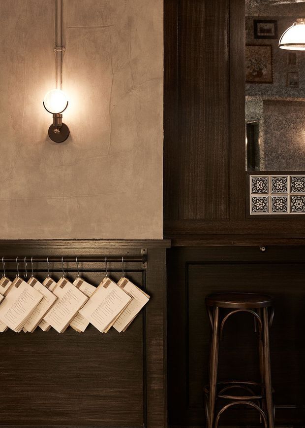 A warmly lit, moody hospitality interior featuring dark timber wall panelling, a textured plaster wall, vintage wall sconces, hanging menus, a decorative tiled panel, framed artwork, and a dark wooden bar stool. The composition suggests a refined bar or restaurant setting with strong traditional detailing and rich brown finishes.